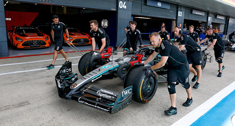 Varios integrantes del equipo empujan un auto de carreras de Fórmula 1 Mercedes-AMG PETRONAS fuera del pit lane, con dos autos naranjas Mercedes-AMG GT en el fondo.