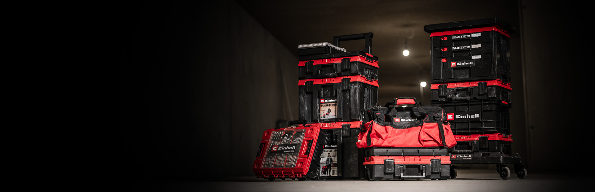 Several black and red Einhell E-Case toolboxes and bags are stacked and arranged in a dark room.