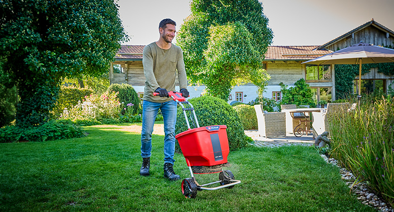 Man pushes red hand spreader with black handle across green lawn in a well-kept garden in front of a house.