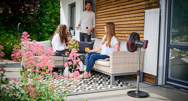 Three people sit on a patio next to a black pedestal fan with a red guard grille and have drinks.