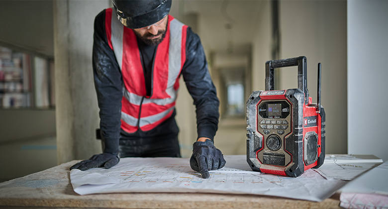 Construction worker wearing a helmet and safety vest pointing at a blueprint, with a red jobsite radio beside him.