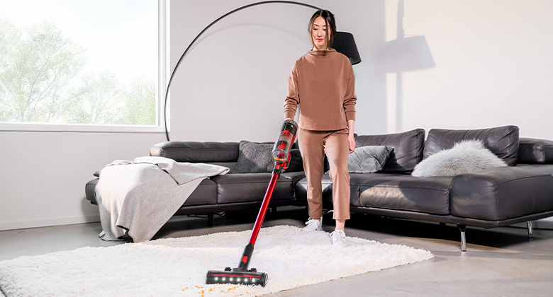 Woman vacuuming a light-colored carpet in the living room with a red cordless vacuum in front of a gray sofa.