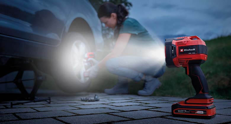 An Einhell battery lamp sheds light on a woman changing a car tyre in the twilight.